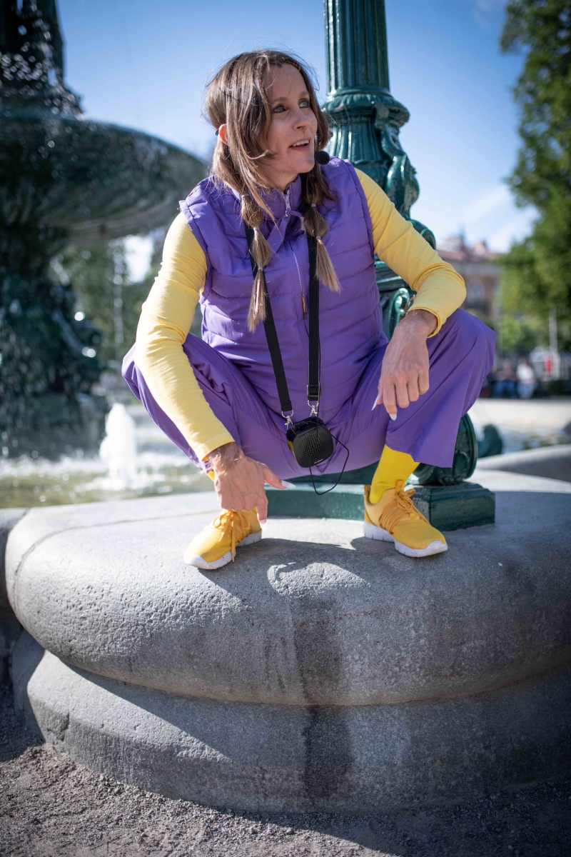 Guide in purple and yellos clothing, long braids and hairy arms crouching on the foot of a fountain, pointing at an old peeing spot.